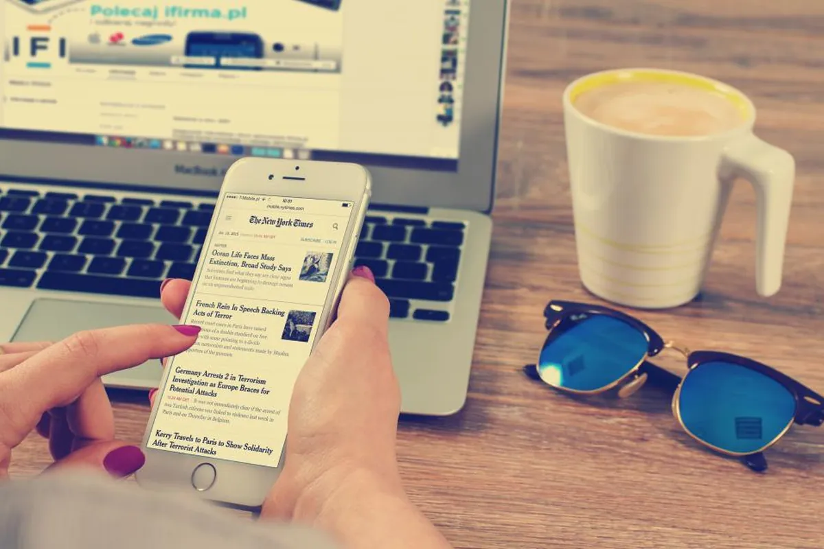 Hands holding a smartphone showing a news article beside a laptop and coffee