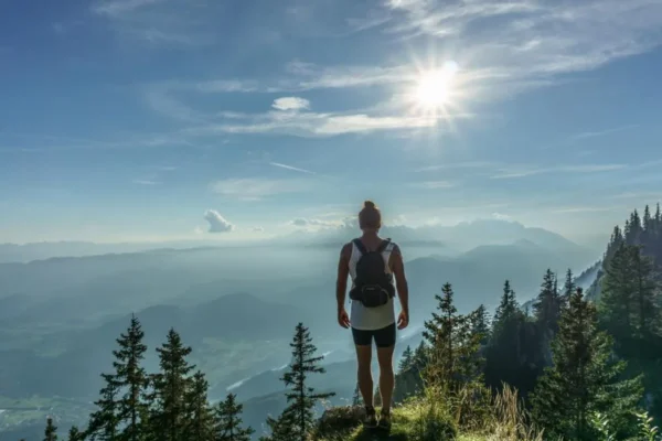 Slow Travel: Why More People Are Choosing Fewer, Deeper Journeys Backpacker standing on a hilltop overlooking forested mountains and a sunlit valley